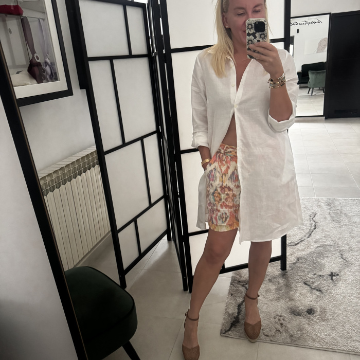 Žena nosi bijelu lanenu košulju dužeg kroja preko šarenih kraktih hlačica